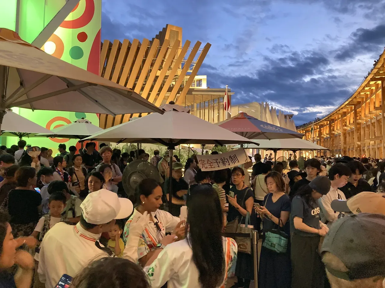 夕暮れの万博会場。パラソルの並ぶ飲食エリアに来場者があふれ、奥に大屋根リングが照らされている
