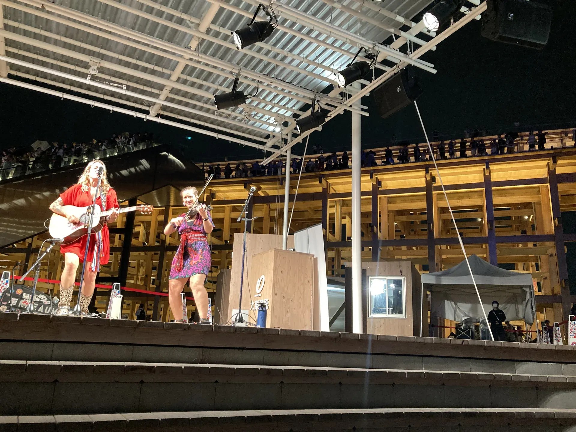 カナダナショナルデーの夜、光の広場〈雲のステージ〉で演奏するカナダ人アーティストのギター＆バイオリン・デュオ、背後で照明に温まる大屋根リングの木組み