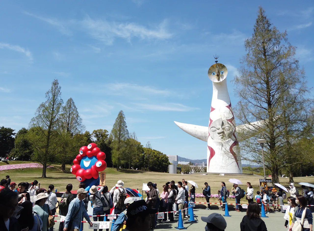 万博記念公園の太陽の塔とミャクミャク像の前に、開幕1周年イベントの来場者が大勢集まっている