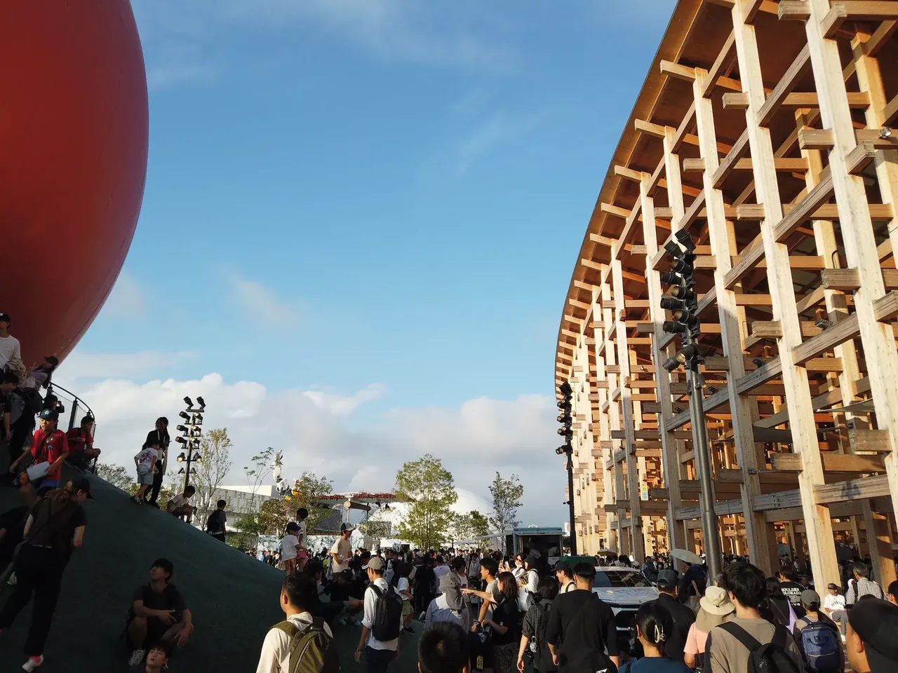夕方の万博会場。木造の大屋根リングを見上げる位置に人があふれ、左手の丘には登って景色を眺める人たちがいる