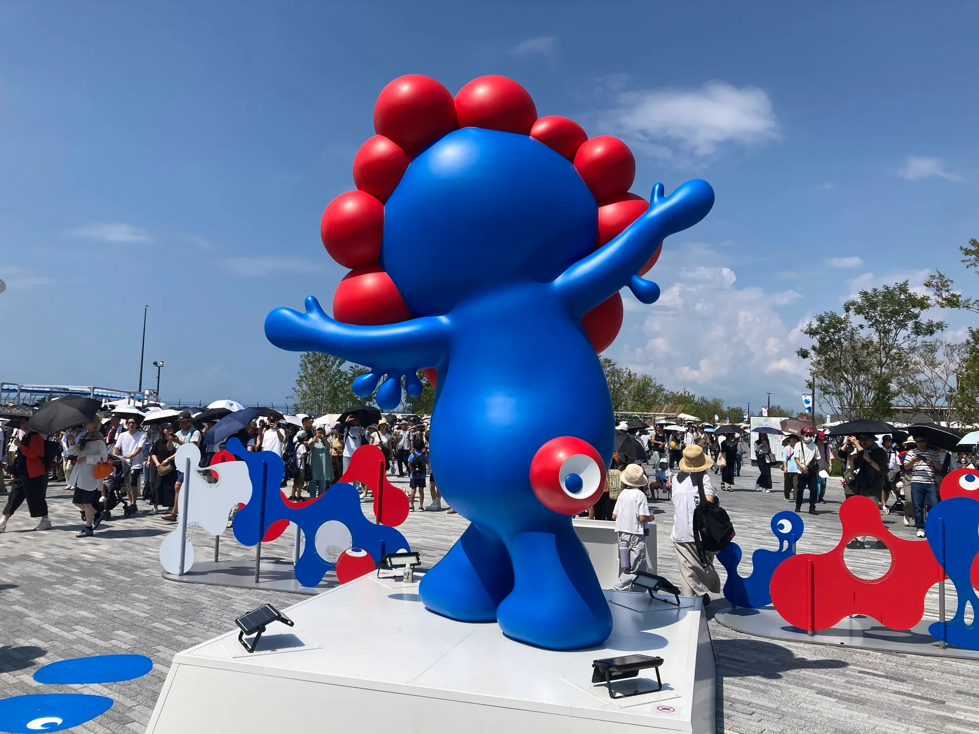 夏空の下、両手を広げたミャクミャク像を日傘の来場者が取り囲み、順番に写真を撮っている