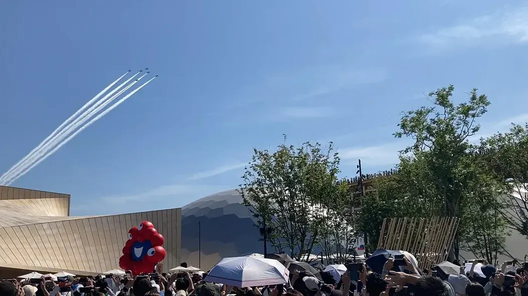 夢洲の会場上空を編隊飛行するブルーインパルスと、見上げる大勢の来場者、ミャクミャクのバルーン
