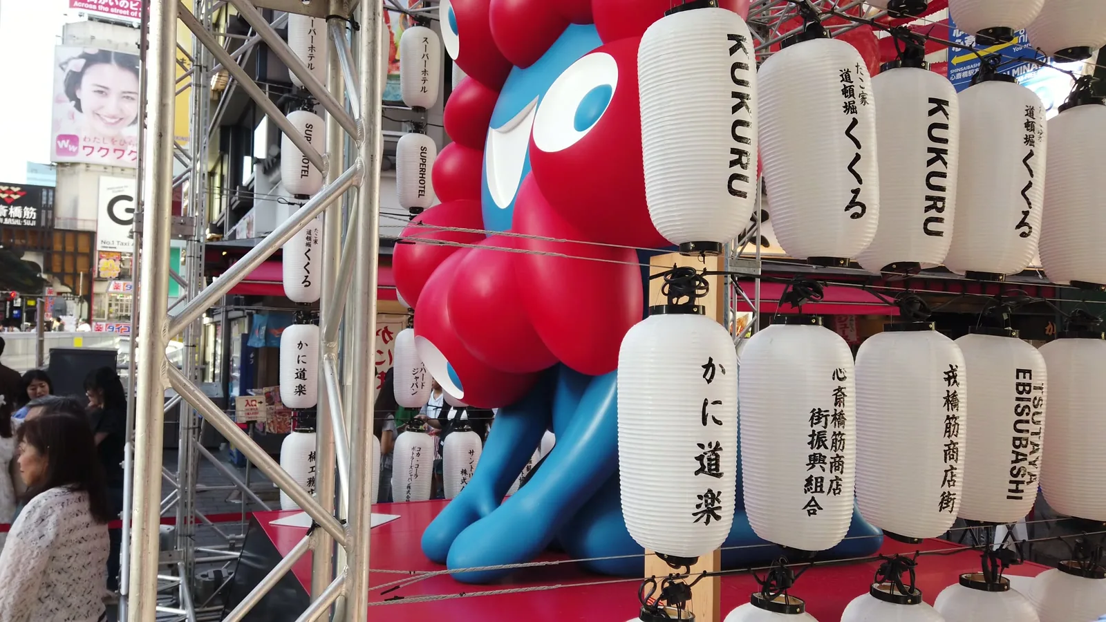祭りの提灯越しに見あげたミャクミャクの像。提灯には戎橋筋商店街や心斎橋筋商店街の名が並ぶ