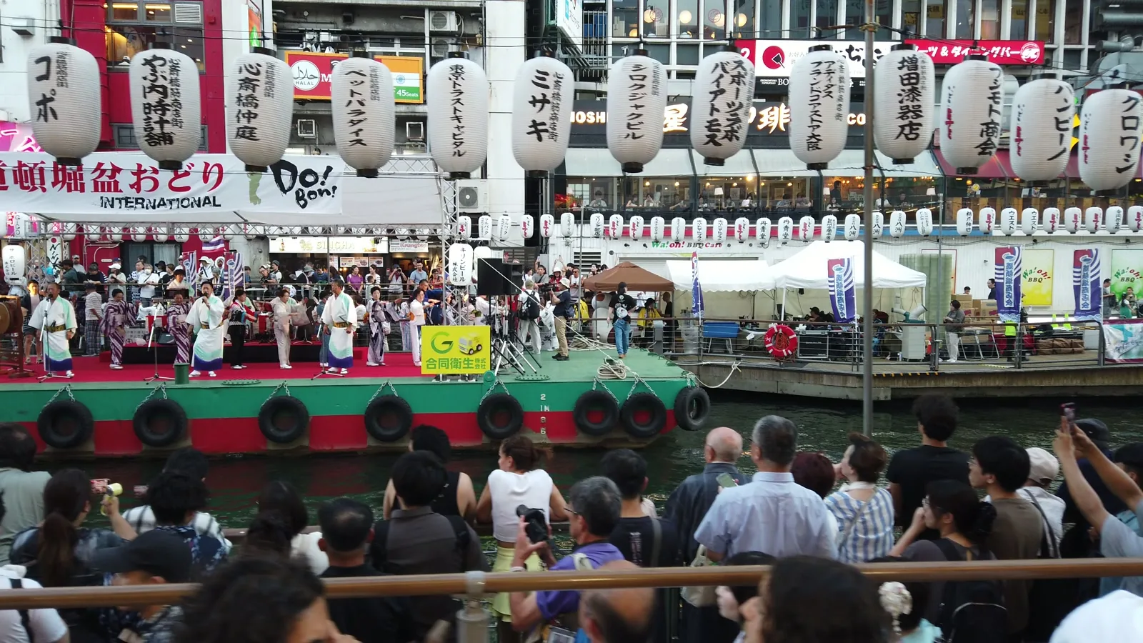 道頓堀川に係留された船の上のステージで踊る浴衣姿の人たちと、対岸の遊歩道を埋める見物客