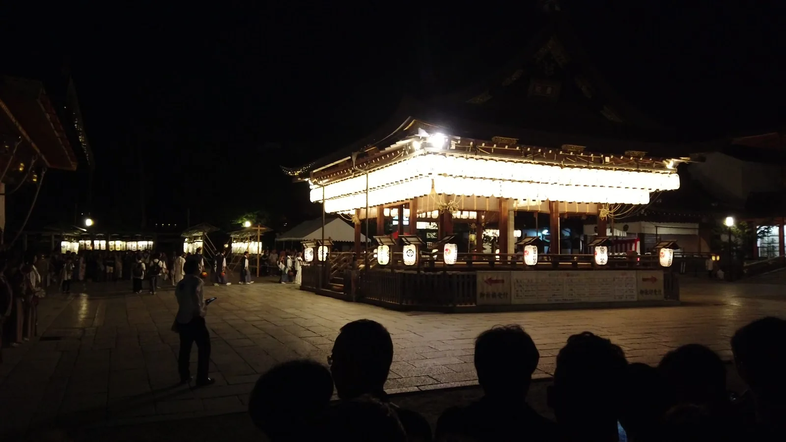 夜の八坂神社。提灯にすべて灯が入って光る舞殿と、その前で神輿を待つ人々のシルエット
