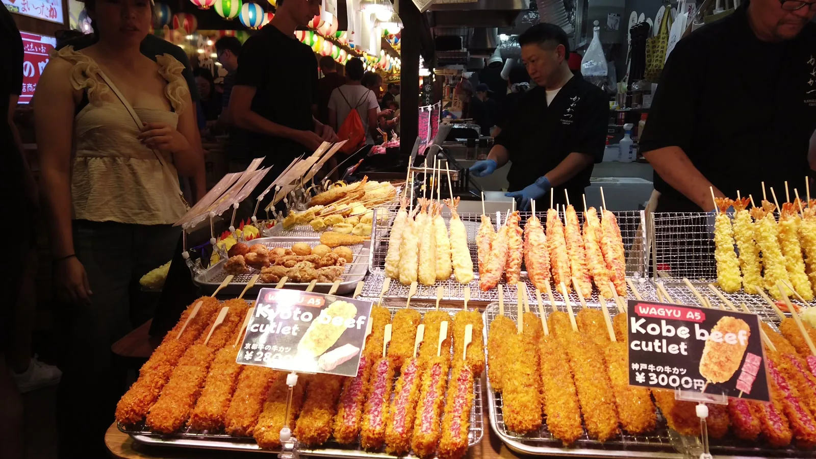 錦市場の店先に並ぶ串カツやえび天。「Kyoto beef cutlet」「Kobe beef cutlet」の英語の値札が立っている