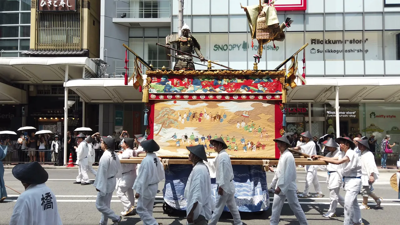 現代的な商業ビルの前を、懸装品をかけた山鉾がゆっくりと通過していく
