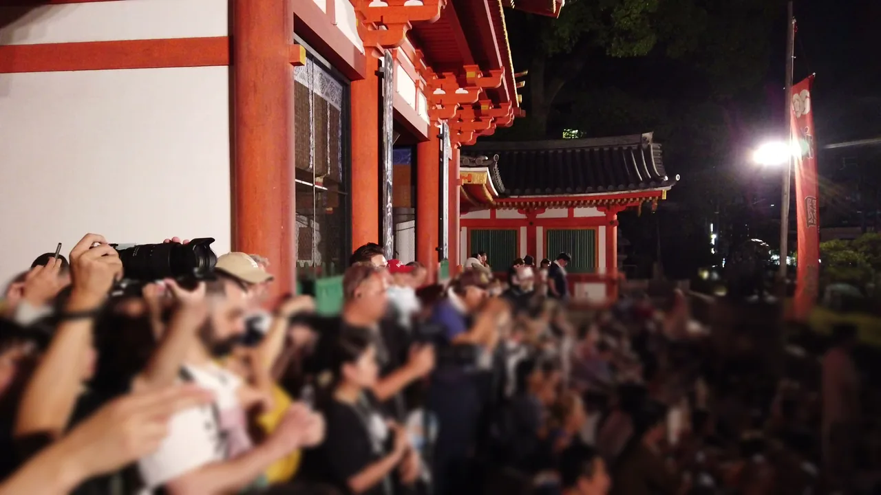 夜の八坂神社境内で、カメラやスマートフォンを掲げて神輿の到着を待つ人々