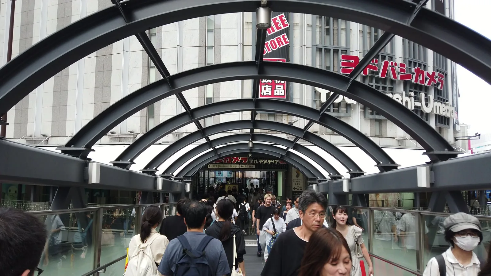 アーチ型の屋根がかかった連絡橋を歩く人たち。正面にヨドバシカメラの看板が見える