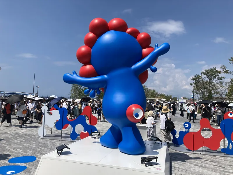 両手を大きく広げたミャクミャク像を背面から見たところ。まわりを日傘の来場者が歩いている