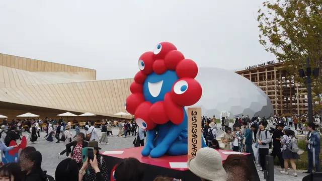 大屋根リングとシェル型パビリオンを背に立つミャクミャク像と来場者