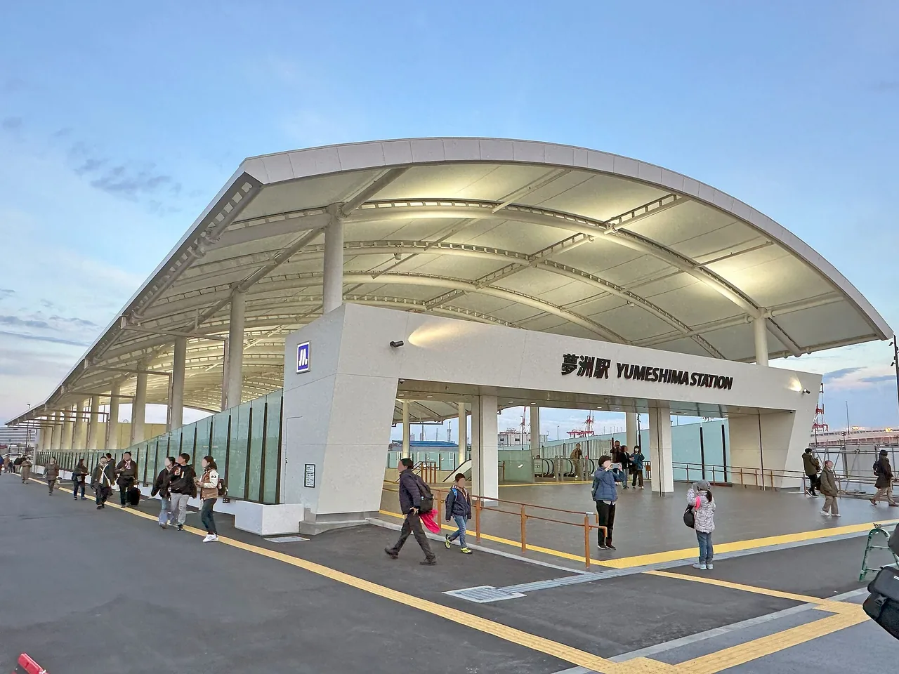 夕暮れの夢洲駅の外観。大きな曲面の屋根の下に「夢洲駅 YUMESHIMA STATION」の表示がある